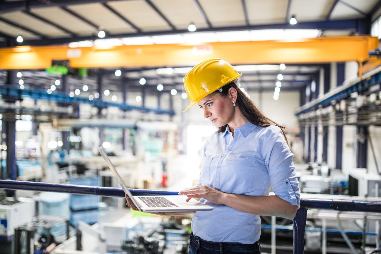 A woman in a hard hat in a warehouse checks her laptop