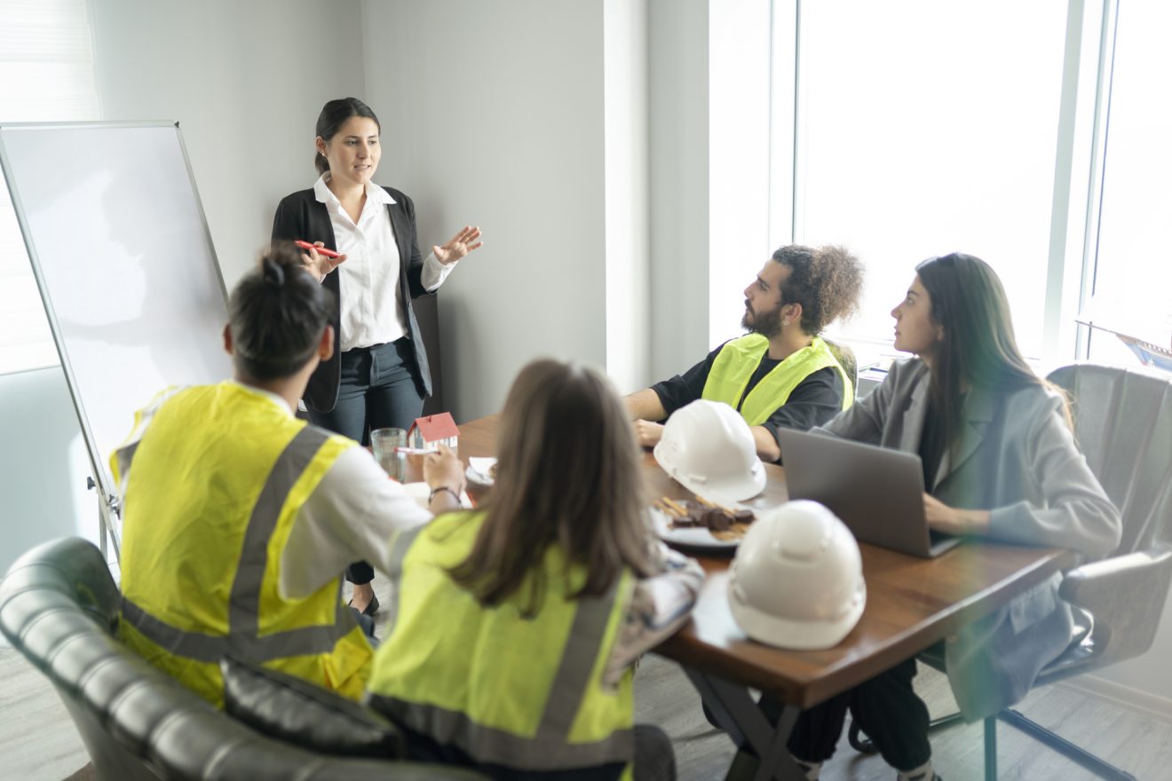 Group of people in bright safety vests with hard hats on the table, listening to a team leader in a business suit