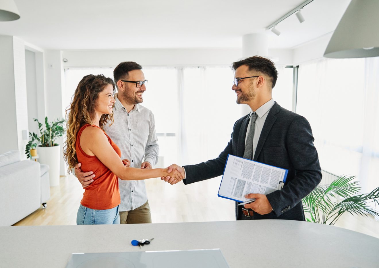 Man in suit holding a business contract, shaking hands with a woman who is standing next to man, who has his hand around her