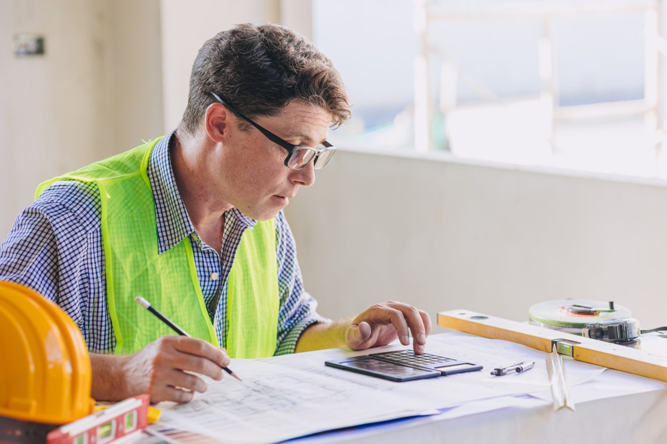 A person in a safety vest reviews schedules and uses a calculator at a desk.
