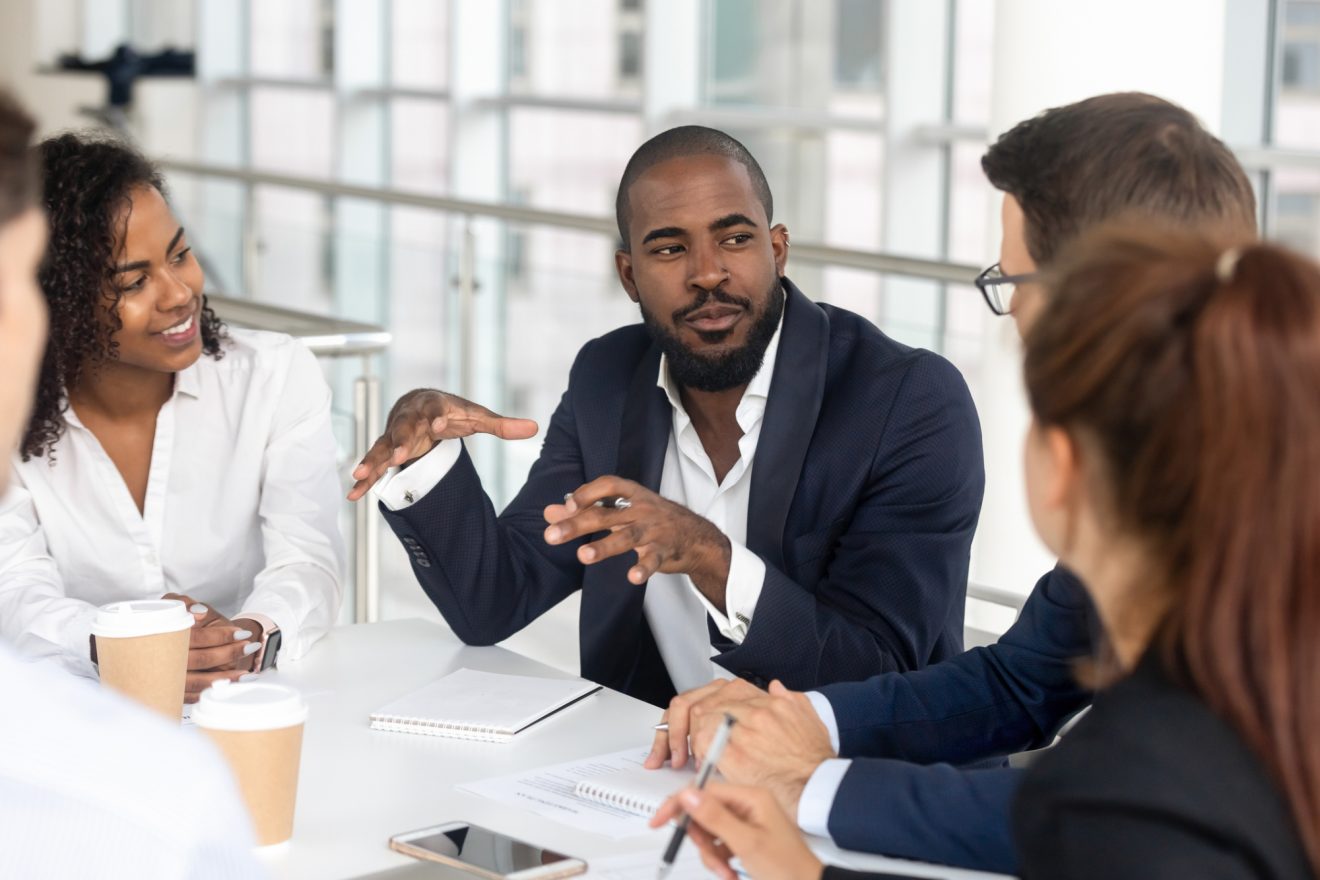 Group of people at a desk listening to a man talk