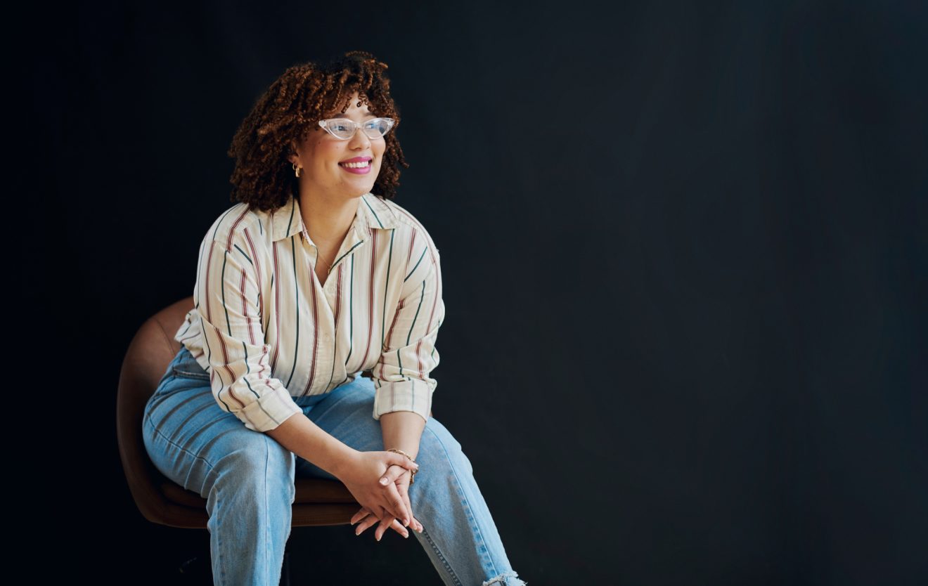 A woman sits in a chair against a dark backdrop and smiles, looking at an angle beyond the camera