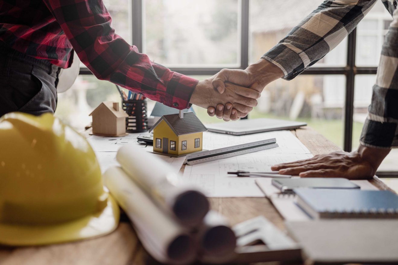 Two men at a construction site shaking hands across a table covered with building models, blueprints, and a hard hat