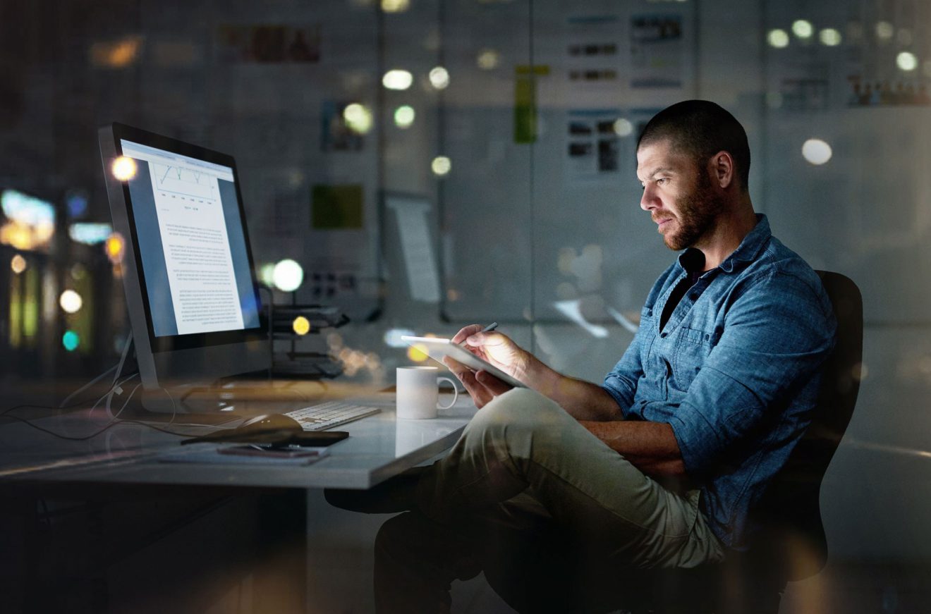 A man sits at a desk in front of a computer in a nighttime interior office scene.