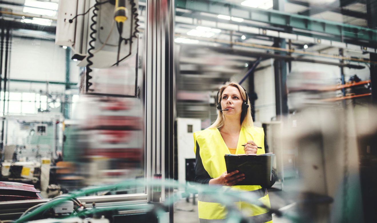 A woman holding a clipboard and wearing a headset in a safety vest observes machinery.