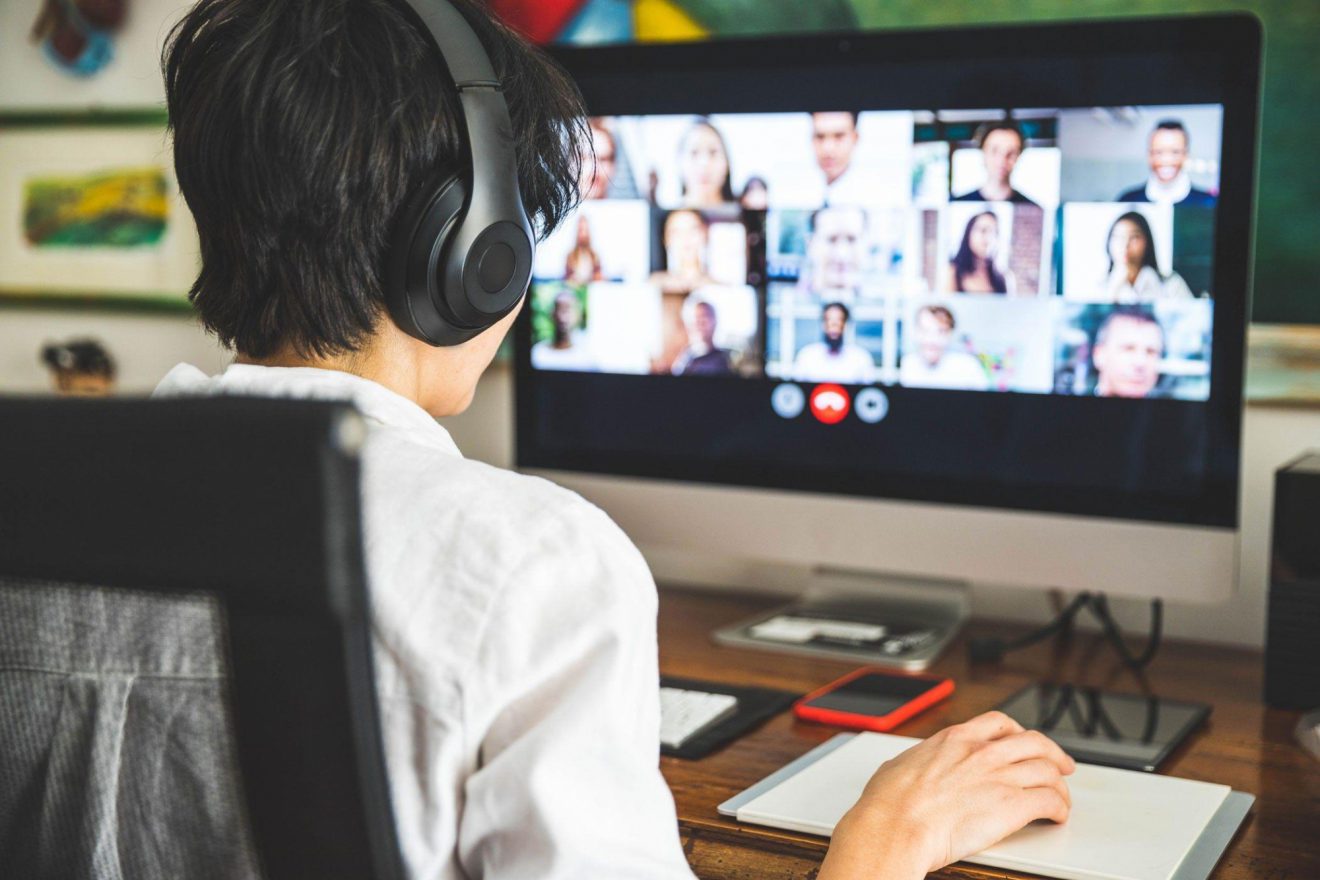 A person wearing headphones sits at a desk in front of a computer screen showing many participants in an online meeting.