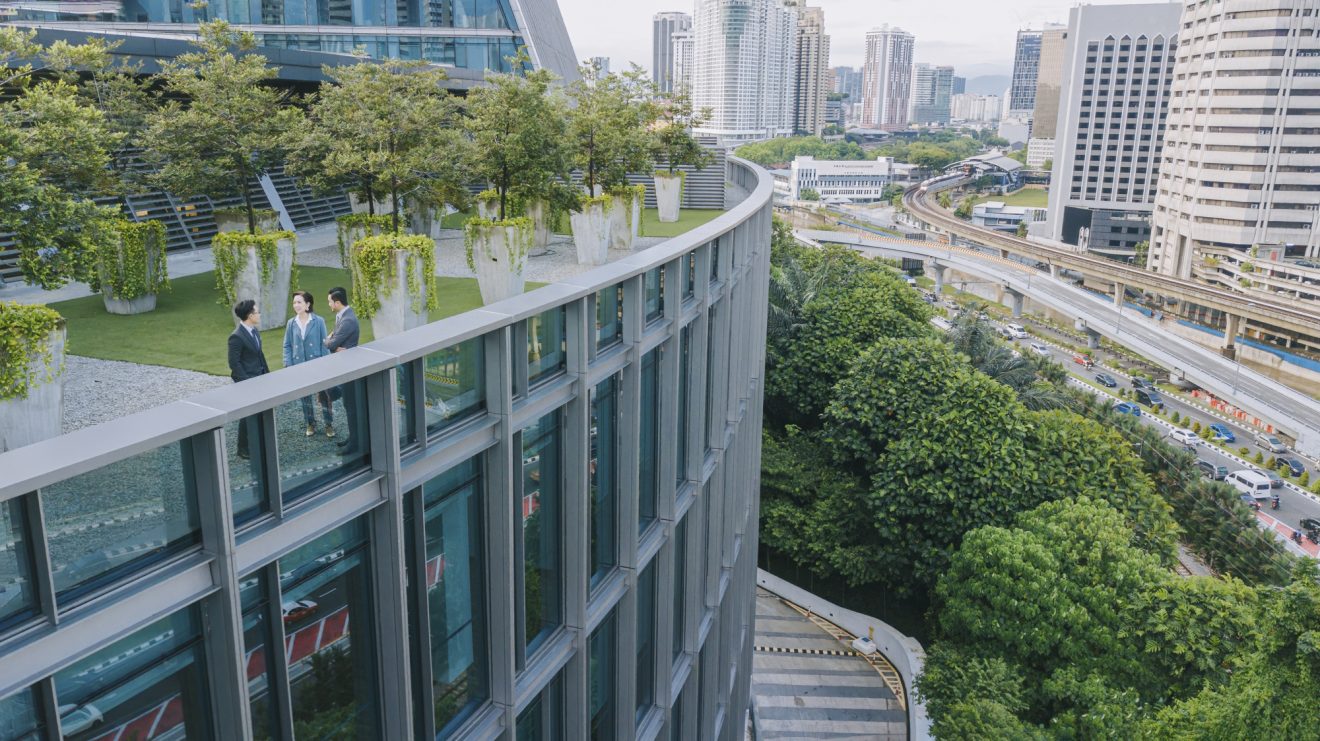 Three people talk on the balcony of a large, curved building surrounded by greenery