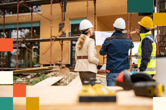 two men and woman in hardhats look over construction plan while looking at building under construction
