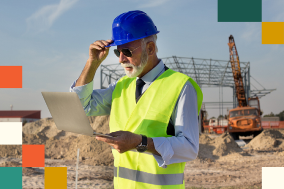 older man wearing a hard hat and reflective vest looking at tablet computer with construction crane in the background