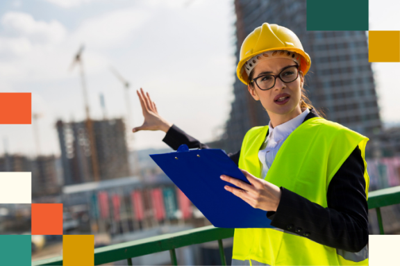 female construction manager wearing a heard hat and holding a clipboard pointing to a building under construction