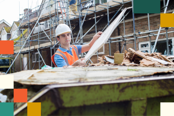A man in a hard hat and reflective vest places wood boards into a dumpster
