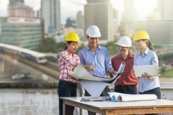 2 women and 2 men in hard hats stand at a table looking at a set of blueprints with a city skyline and train in the background